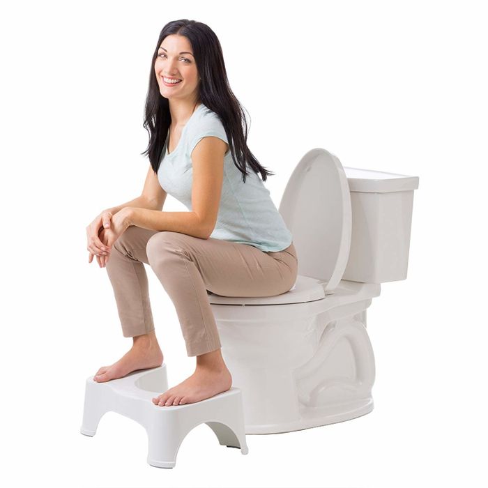 A fully dressed model sits on the toilet with their feet resting on the stool, knees raised above their hips