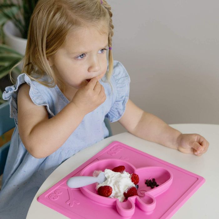 A small child eating from a pink silicone Peppa Pig mat with distinct sections for food, placed on a table