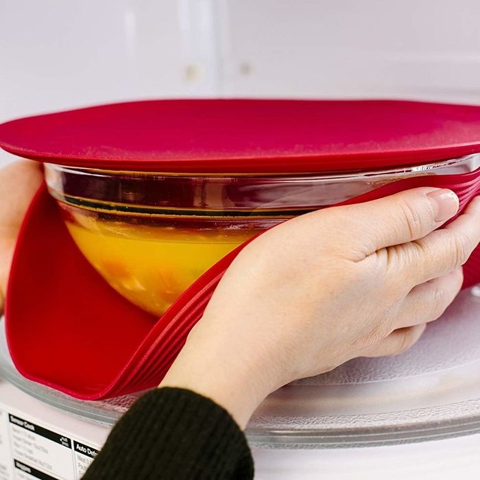 Two hands gripping both sides of a bowl covered with the small silicone lid while the larger cover rests nearby