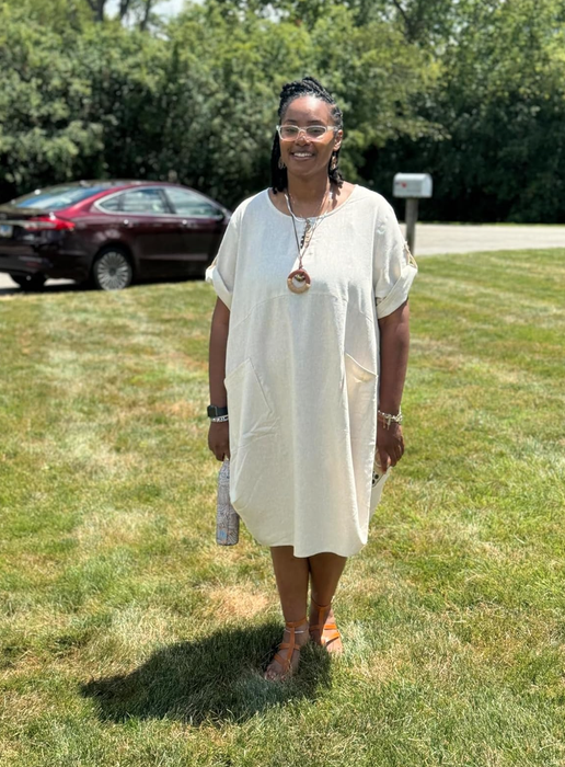 A woman in a light-colored, loose-fitting dress stands on grass, with trees and a car in the background
