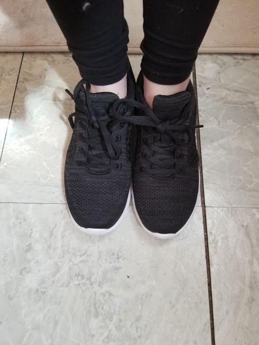A person wearing black athletic sneakers with white soles, standing on a tiled floor