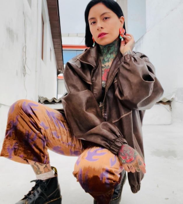 A person with colorful tattoos kneels, wearing a loose-fitting jacket, velvet pants, and black boots, with dark hair and bold earrings.
