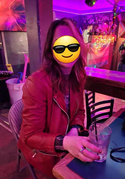A person, with a smiling face emoji covering their face, rocks a fashionable leather jacket while sipping on a drink and sitting at a bar.