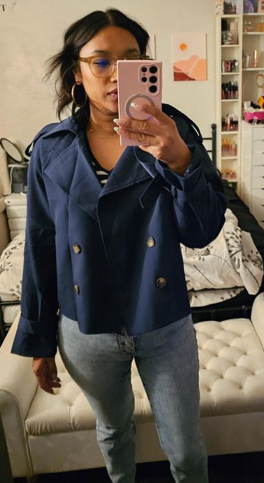 A person posing in front of a mirror, wearing a double-breasted blazer, jeans, and glasses, taking a selfie with a pink phone in what looks like a bedroom.
