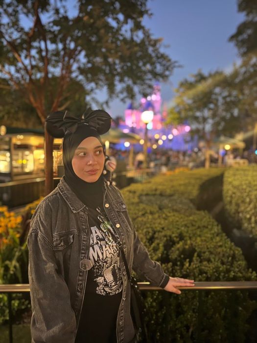 A person wearing a Mickey Mouse ears headband and a casual denim jacket stands by trimmed hedges at an outdoor shopping area, with an illuminated castle softly blurred in the background.