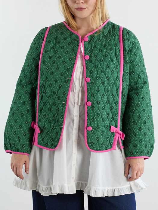 A woman models a green quilted jacket with pink accents, layered over a white ruffled blouse and blue pants, giving off a playful yet chic vibe.