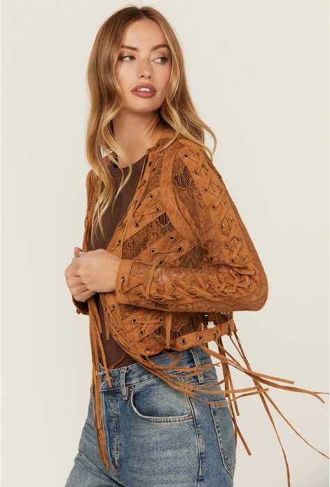 A woman wears a fringed, brown leather jacket over a brown top and jeans. The jacket showcases intricate lace-up details.
