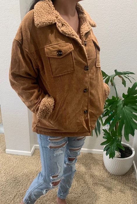 A person models a sherpa-lined corduroy jacket paired with distressed denim jeans, standing next to a potted plant.