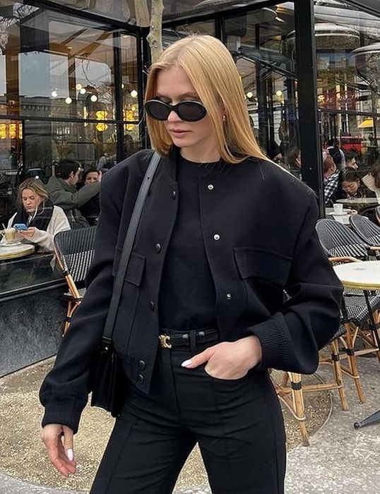 A woman in a black jacket, black pants, and sunglasses, carrying a black shoulder bag, standing in an outdoor café setting.