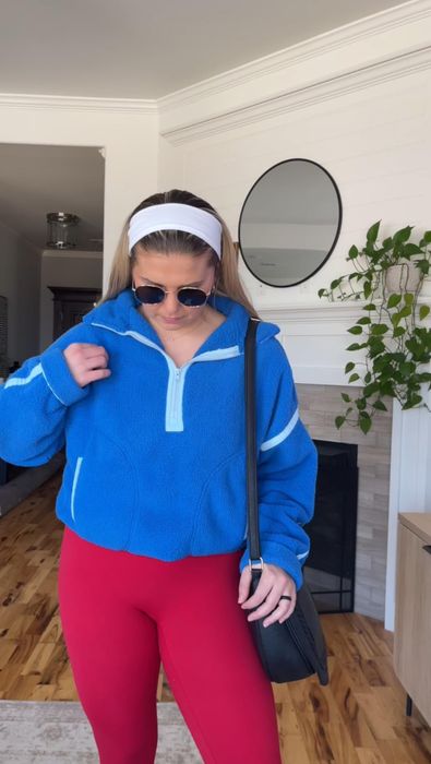 A woman dons a cozy blue fleece, red leggings, sunglasses, and a white headband, standing in a modern living room adorned with plants and a circular mirror.