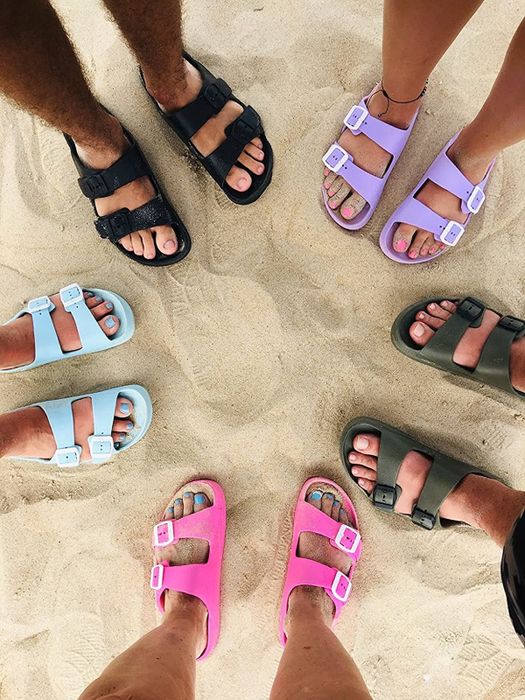 A reviewer photo showing people wearing the slides in pink, light blue, purple, and dark green as they walk along the beach.