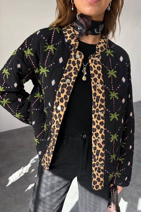 A person is casually posing in a patterned jacket adorned with leopard and palm tree motifs, a black shirt underneath, and a scarf wrapped around their neck.