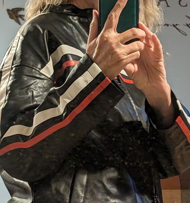 A person takes a selfie in front of a mirror, focusing on their leather jacket with bold black, white, and red stripes on the sleeves.
