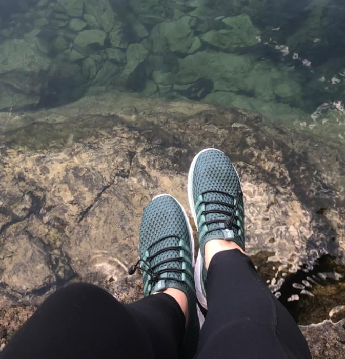 A reviewer sporting gray-green sneakers while standing near a body of water