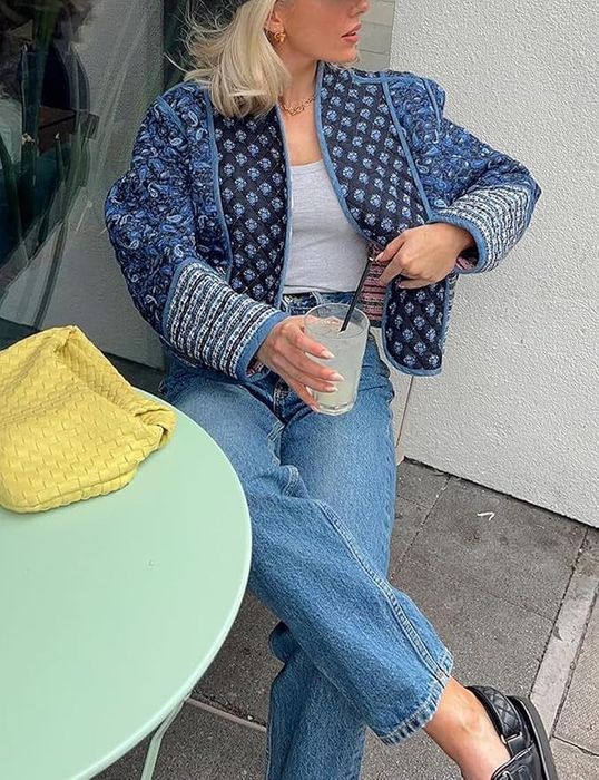 A blonde woman dressed in a patterned jacket, white shirt, and blue jeans, enjoying a drink, with a yellow bag placed on a green table outside.