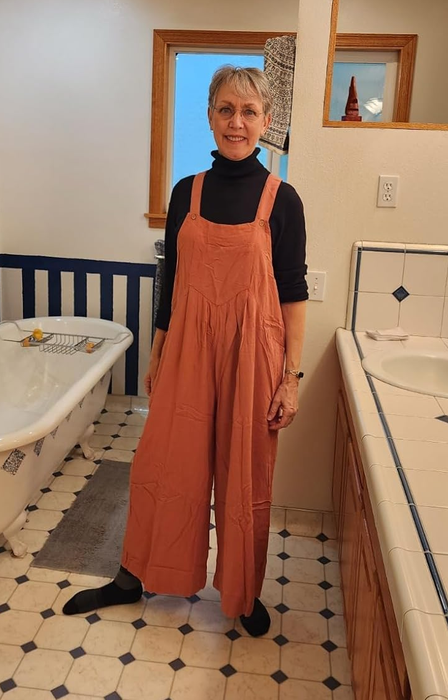 A reviewer strikes a pose in an orange jumpsuit worn over a black long-sleeve top.