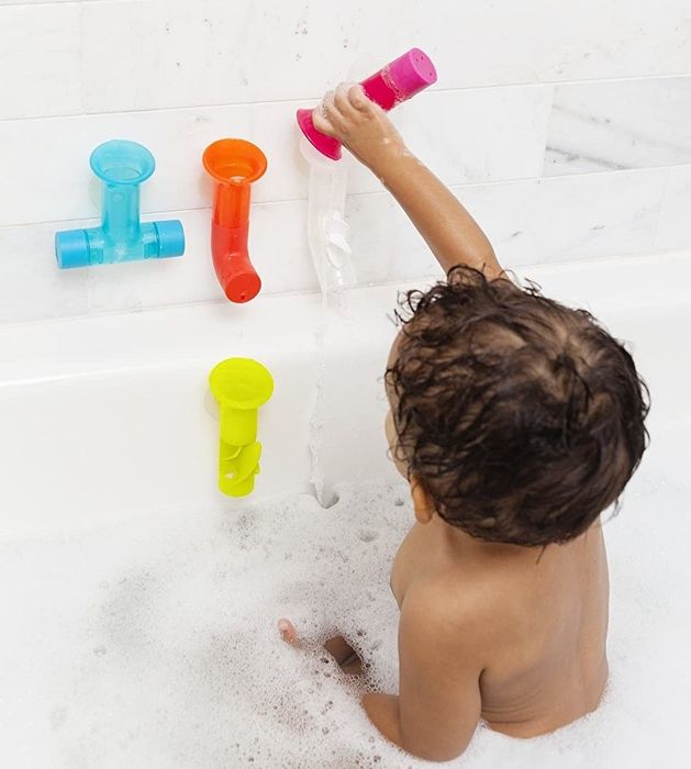 A baby sitting up, pouring water through pipes attached to the bathtub wall