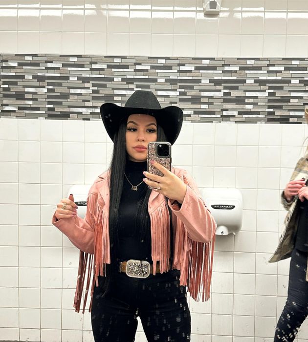 A woman takes a mirror selfie in a bathroom, dressed in a black hat, black turtleneck, a pink fringed jacket, and a belt with a large buckle. Another person is partially visible in the shot.