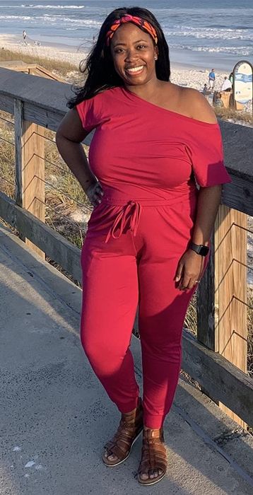 A reviewer smiling while wearing a dark red off-shoulder jumpsuit on a beachside boardwalk.