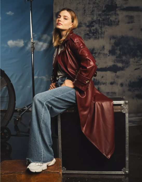 A woman in a long leather trench coat and wide-leg jeans poses atop a crate in a studio, with photography equipment visible in the background.