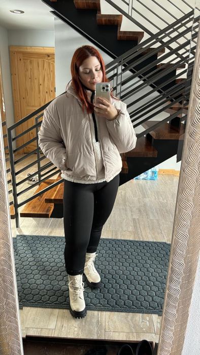 A woman strikes a pose in front of a sleek staircase, wearing a puffy jacket, black leggings, and fur-lined boots while snapping a mirror selfie.