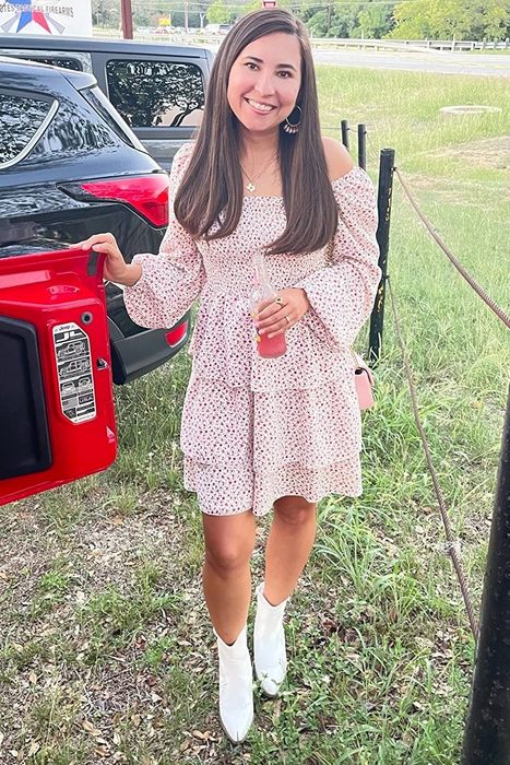 A reviewer stands next to a parked car, wearing a white off-shoulder floral dress and white boots, holding a drink in a grassy area.