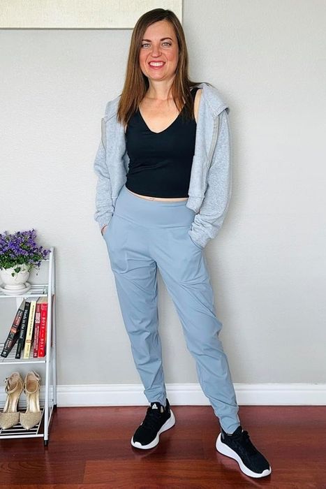 A reviewer wearing a black top, gray jacket, light blue pants, and black sneakers standing indoors.