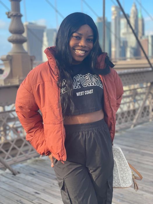 A woman smiles on a city bridge, dressed in a West Coast cropped top, puffer jacket, and joggers, holding a chic white handbag.