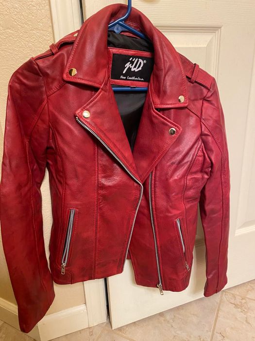 A red faux leather jacket with silver zippers and button details, hanging neatly on a blue hanger in front of a white door.