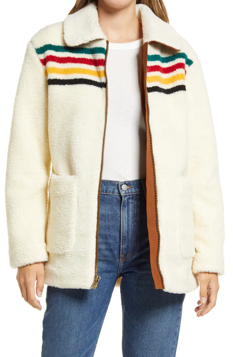 A model is seen wearing a cream-colored sherpa jacket with colorful stripes across the chest, paired with a white shirt and blue jeans.