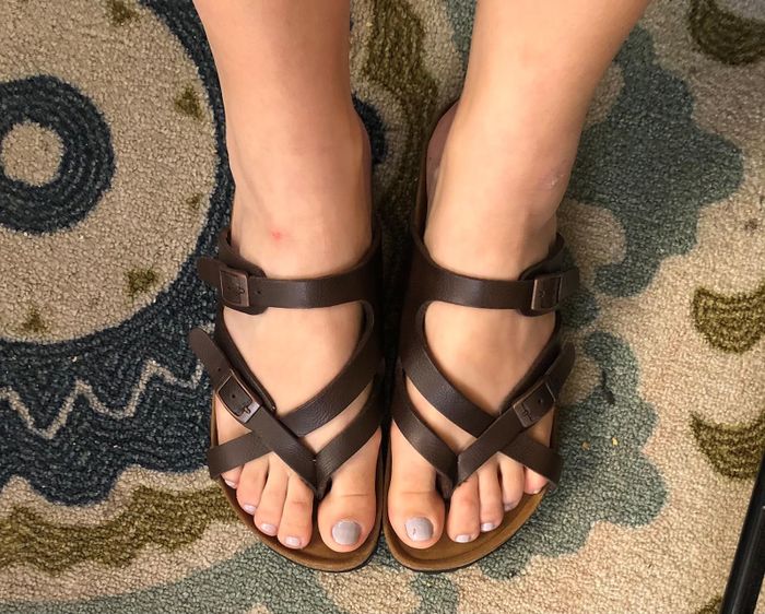 A close-up of a reviewer’s feet wearing brown strappy sandals on a patterned rug