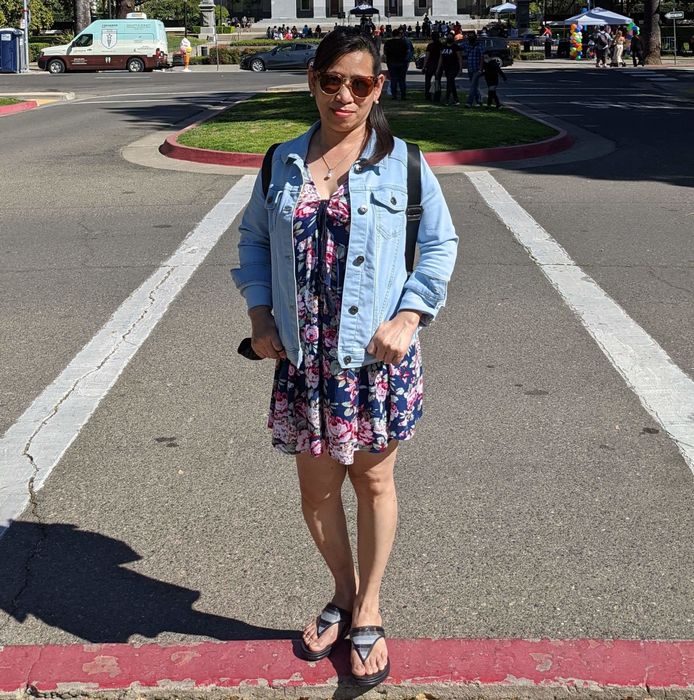 A reviewer wearing a light blue jacket over a dress, standing in front of a legislative building.
