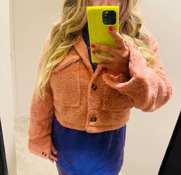 A person snaps a mirror selfie, wearing a fluffy, buttoned jacket paired with a blouse or dress, holding a phone with a yellow case.