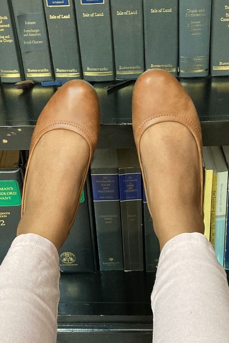 A reviewer sitting with their feet resting on a shelf of books, wearing tan flats