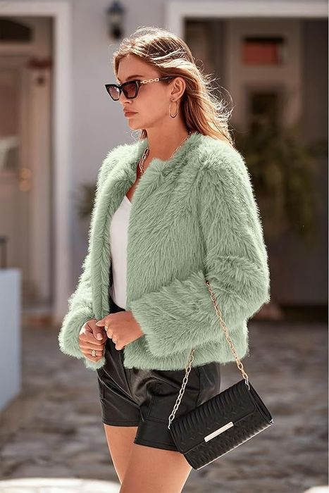 A woman walks outdoors, rocking a fuzzy jacket, white top, black shorts, sunglasses, and a black purse with a gold chain, her medium-length hair flowing.
