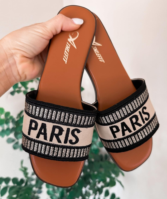 Hand holding a pair of beige and black sandals with the word