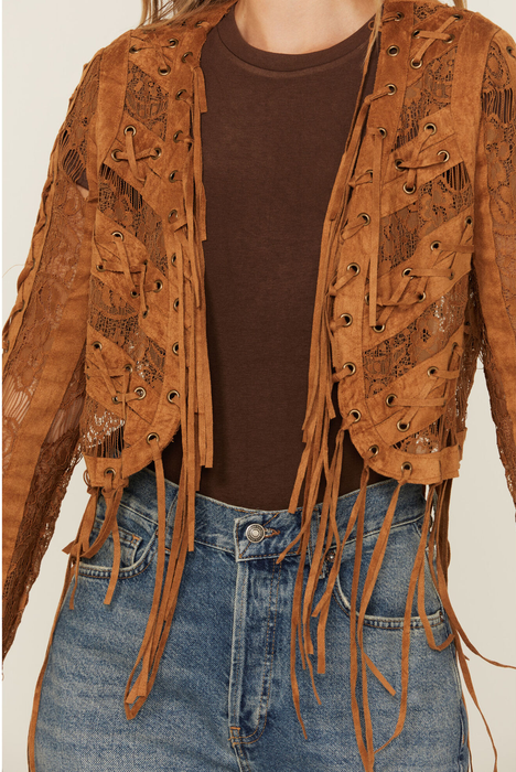 A woman is dressed in a brown fringed and lace-up suede jacket, layered over a brown top and blue jeans.