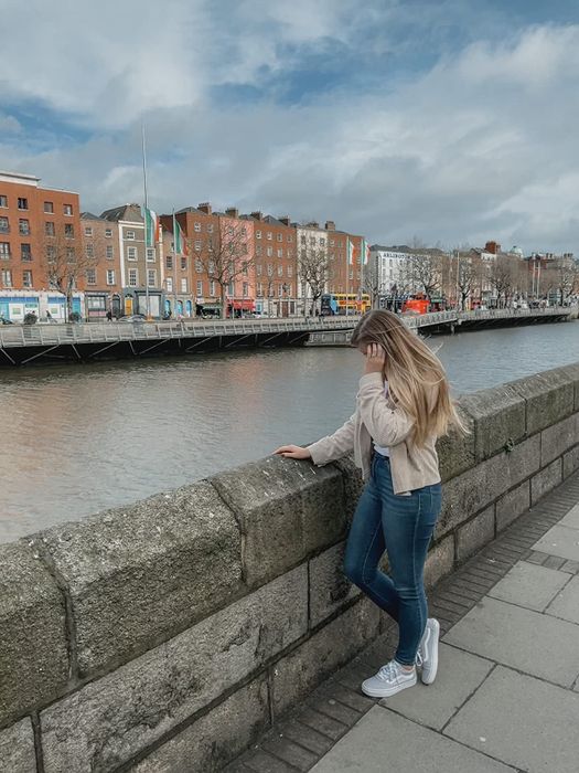 A woman with long hair, dressed casually in jeans and a beige jacket, stands near a stone railing by the river, gazing at the peaceful water.