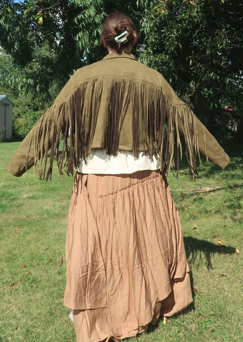 A person stands outdoors, wearing a tan tattered skirt, cream blouse, and fringed jacket, with their long hair pinned up in a clip.