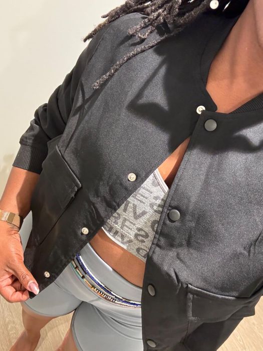 A person dons a black button-up jacket over a gray sports bra with visible text, matched with light gray athletic shorts that have a beaded waistband.