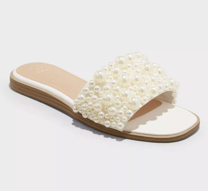 A white slide sandal featuring a flat sole and a strap adorned with several large pearls for a stylish touch