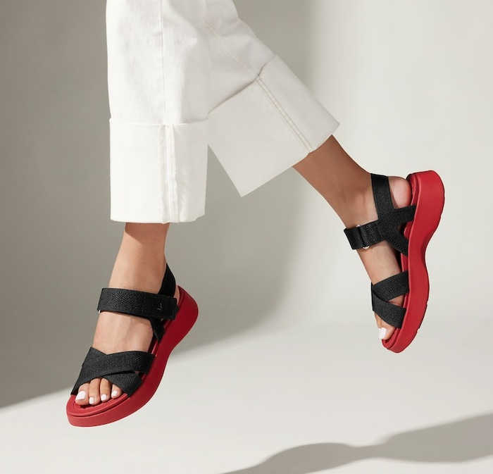 A person wearing white pants, shown mid-air, highlighting black and red sandals with thick straps, with the focus on the shoes to suggest a shopping context
