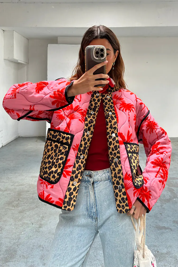 A person snaps a mirror selfie, sporting a jacket featuring a blend of floral and leopard prints, a red top, and light blue jeans, with a white handbag in hand.