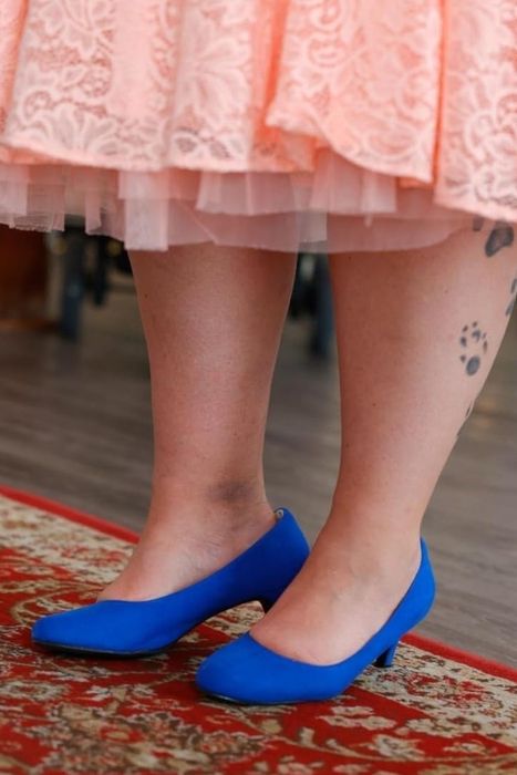 A reviewer in a peach lace dress, wearing bright blue pumps, standing on an ornate rug