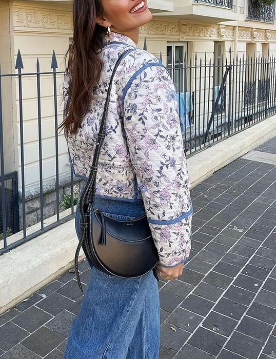 A woman, seen from behind, walks along a sidewalk near a wrought iron fence, sporting a chic floral jacket and jeans, and carrying a black leather shoulder bag.