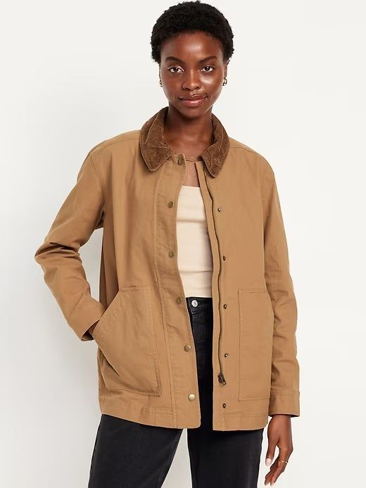 A woman dressed in a casual tan jacket over a beige top and black jeans, featuring a button-down front and brown collar trim.