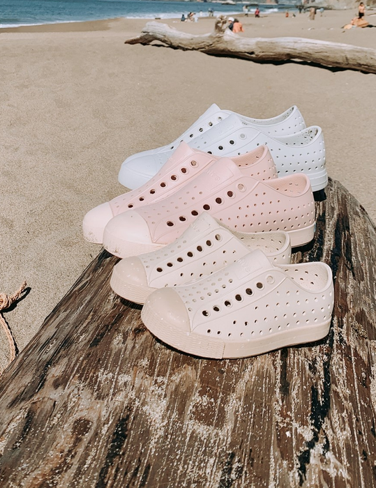 several pairs of Jefferson Bloom sneakers placed on a log at the beach, available in three colors: white, light pink, and beige