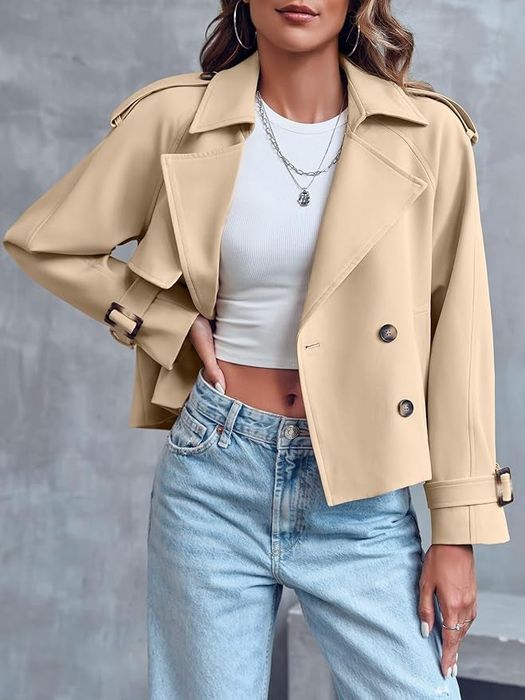 A woman sporting a beige trench coat over a white crop top and high-waisted jeans, accessorized with layered necklaces. This look is set in a shopping context.