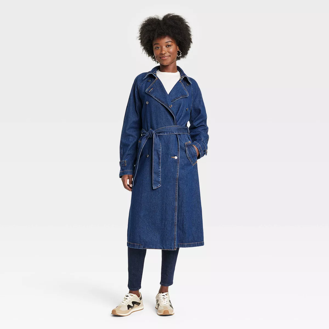 A woman models a denim trench coat with a belted waist, paired with a white top, dark pants, and sneakers, smiling as she stands.
