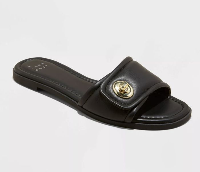 A black, open-toe slip-on sandal featuring a gold emblem on the strap